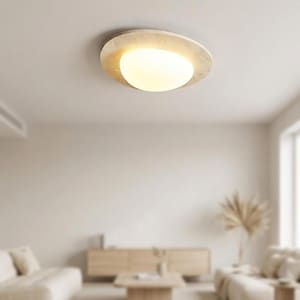 May include: A modern, circular ceiling light fixture with a warm, glowing light. The fixture has a textured, off-white outer ring and a rounded, frosted glass center. The light is mounted on a white ceiling, and the room has a neutral color palette.