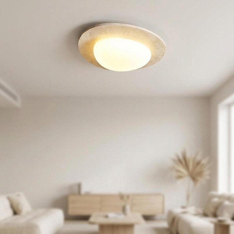 May include: A modern, circular ceiling light fixture with a warm, glowing light. The fixture has a textured, off-white outer ring and a rounded, frosted glass center. The light is mounted on a white ceiling, and the room has a neutral color palette.