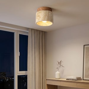 May include: A ceiling-mounted light fixture with a cylindrical, beige stone shade and a wooden top. The light is on, illuminating the interior. The fixture is in a room with a window, curtains, and a wooden console table.