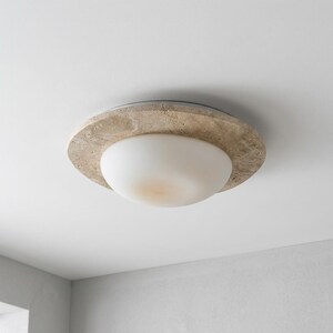 May include: A ceiling light fixture with a circular, textured, beige stone-like outer ring. The light has a white, rounded, glass-like shade. The fixture is mounted on a white ceiling.