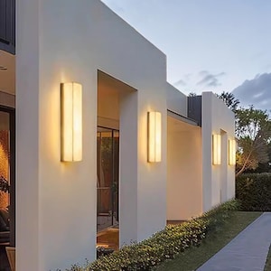 May include: Exterior view of a modern building with several rectangular wall-mounted lights. The lights have a warm, inviting glow and are mounted on a white exterior. A pathway and greenery are visible in the foreground.