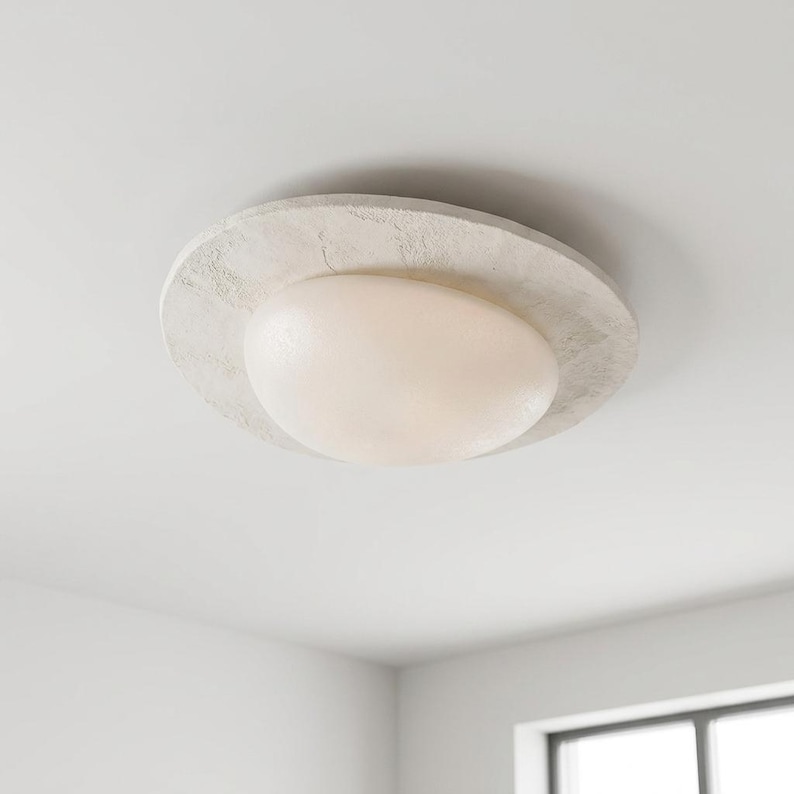 May include: A modern ceiling light fixture with a textured, off-white circular base and a rounded, opaque, cream-colored light. The fixture is mounted on a white ceiling, providing ambient illumination for a room.