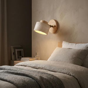 Può includere: Una lampada da parete con un paralume strutturato di colore chiaro e una base in legno. La lampada è posizionata sopra un letto con biancheria da letto dai toni neutri. La calda luce della lampada illumina la stanza, creando un'atmosfera accogliente.