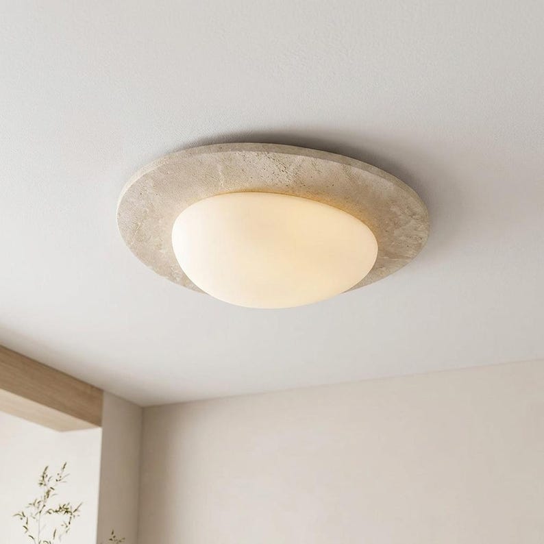May include: A modern ceiling light fixture with a round, textured, light gray concrete-like frame. The light has a large, oval-shaped, frosted glass shade that emits a soft, warm glow. The fixture is mounted on a white ceiling.