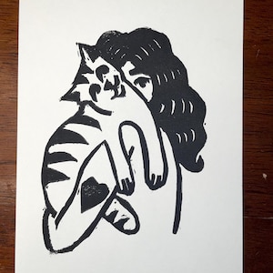 Puede incluir: Linograbado en blanco y negro de una persona sosteniendo un gato. El gato tiene marcas de rayas y una forma de corazón en su costado. El cabello de la persona es largo y ondulado.