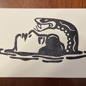 May include: Black and white linocut print depicting a person in water being attacked by a snake. The snake is arched over the person with its mouth open. The print is on a white background.