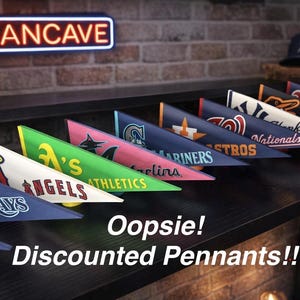 Puede incluir: Estandartes de equipos de béisbol, incluyendo los Angels, Athletics y Mariners, dispuestos sobre una superficie oscura. Un letrero de neón "MANCAVE" es visible en el fondo. El texto "Oopsie! Discounted Pennants!!" también está presente.