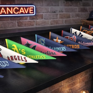 Peut inclure: Une collection de fanions triangulaires avec les logos de diverses équipes de baseball de la Ligue majeure, disposés sur une surface sombre. Un néon "MANCAVE" est visible en arrière-plan, ainsi qu'une balle et une casquette de baseball.