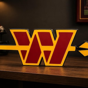 May include: A decorative sign featuring the Washington Commanders logo, a burgundy and gold "W" with an arrow. The sign is on a wooden surface, with a football and helmet in the background. The sign is a sports-themed item.