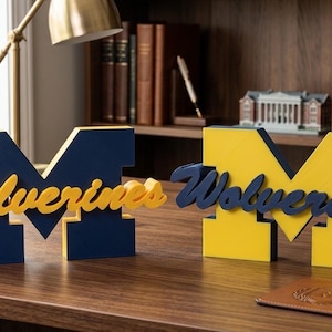 Peut inclure: Deux enseignes en forme de lettre M majuscule avec le mot Wolverines écrit en lettres cursives. L'une est bleu marine avec une inscription jaune, et l'autre est jaune avec une inscription bleu marine. Ces objets décoratifs sont autonomes.