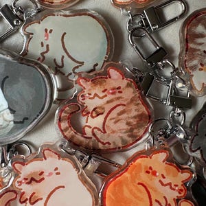 Puede incluir: Una colección de llaveros de gato de acrílico transparente. Cada llavero presenta una ilustración de acuarela diferente de un gato en varias poses y colores, incluyendo naranja, marrón y gris. Los llaveros tienen cierres metálicos plateados.