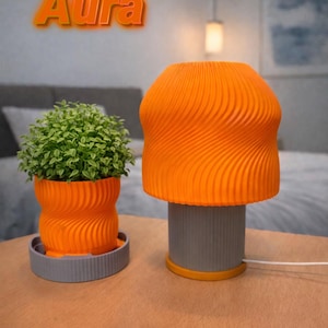 May include: An orange lamp and matching plant pot with a swirling design sit on a wooden table. The lamp has a gray base with an orange top. The plant pot holds a small green plant and sits on a gray tray. The text "The Aura" is in orange above the lamp.