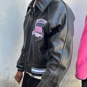 K&ouml;nnte beinhalten: Eine schwarze Lederjacke mit Umlegekragen, Frontrei&szlig;verschluss und einem wei&szlig;-schwarz gestreiften Saum. Auf der Brust befindet sich ein Aufn&auml;her mit dem Text AVIREX &uuml;ber einer US-Flagge und dem Schriftzug ESTABLISHED 1975 darunter.