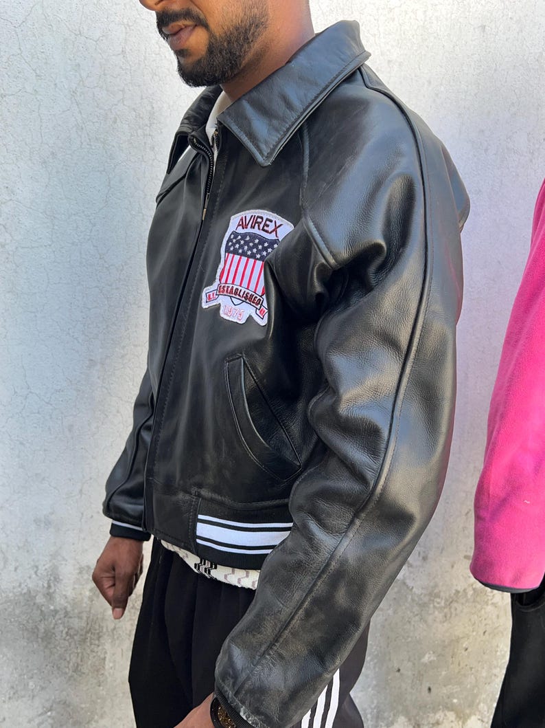 K&ouml;nnte beinhalten: Eine schwarze Lederjacke mit Umlegekragen, Frontrei&szlig;verschluss und einem wei&szlig;-schwarz gestreiften Saum. Auf der Brust befindet sich ein Aufn&auml;her mit dem Text AVIREX &uuml;ber einer US-Flagge und dem Schriftzug ESTABLISHED 1975 darunter.