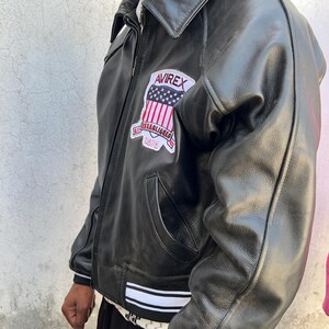 K&ouml;nnte beinhalten: Eine schwarze Lederjacke mit Umlegekragen, Frontrei&szlig;verschluss und einem Aufn&auml;her auf der Brust. Der Aufn&auml;her zeigt den Text AVIREX, ESTABLISHED und 1975 sowie eine US-Flagge. Die Jacke hat gerippte B&uuml;ndchen mit wei&szlig;en Streifen.
