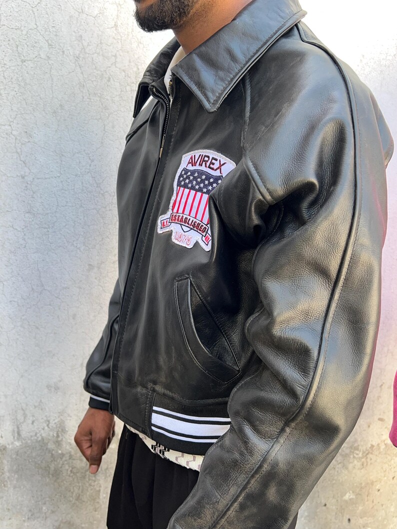 K&ouml;nnte beinhalten: Eine schwarze Lederjacke mit Umlegekragen, Frontrei&szlig;verschluss und einem Aufn&auml;her auf der Brust. Der Aufn&auml;her zeigt den Text AVIREX, ESTABLISHED und 1975 sowie eine US-Flagge. Die Jacke hat gerippte B&uuml;ndchen mit wei&szlig;en Streifen.
