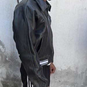 K&ouml;nnte beinhalten: Eine schwarze Lederjacke mit klassischem Kragen, langen &Auml;rmeln und einem Saum, der mit zwei horizontalen wei&szlig;en Streifen verziert ist. Die Jacke verf&uuml;gt &uuml;ber einen Frontrei&szlig;verschluss und Seitentaschen sowie eine glatte Textur.