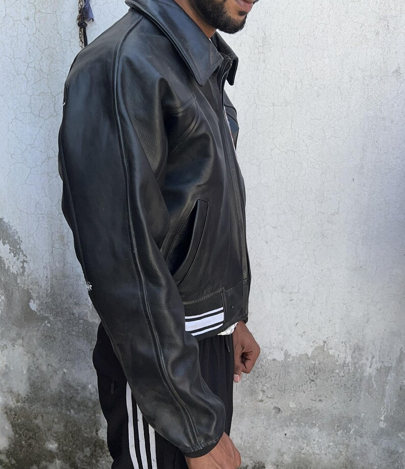 K&ouml;nnte beinhalten: Eine schwarze Lederjacke mit klassischem Kragen, langen &Auml;rmeln und einem Saum, der mit zwei horizontalen wei&szlig;en Streifen verziert ist. Die Jacke verf&uuml;gt &uuml;ber einen Frontrei&szlig;verschluss und Seitentaschen sowie eine glatte Textur.