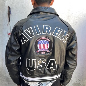 K&ouml;nnte beinhalten: Eine schwarze Lederjacke mit dem Wort AVIREX in gro&szlig;en wei&szlig;en Buchstaben auf dem oberen R&uuml;cken. Darunter befindet sich ein kreisf&ouml;rmiger Aufn&auml;her mit dem Text ALL AMERICANS ESTABLISHED 1975 und einer US-Flagge. Das Wort USA steht in gro&szlig;en Buchstaben.