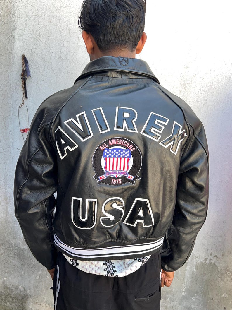 K&ouml;nnte beinhalten: Eine schwarze Lederjacke mit dem Wort AVIREX in gro&szlig;en wei&szlig;en Buchstaben auf dem oberen R&uuml;cken. Darunter befindet sich ein kreisf&ouml;rmiger Aufn&auml;her mit dem Text ALL AMERICANS ESTABLISHED 1975 und einer US-Flagge. Das Wort USA steht in gro&szlig;en Buchstaben.