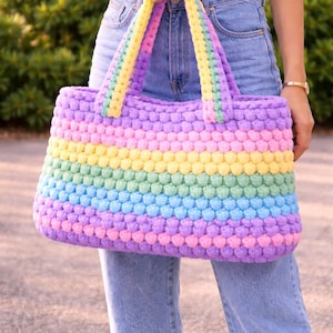 May include: A colorful, handmade crochet tote bag. The bag features horizontal stripes in pastel shades of pink, yellow, green, blue, and purple. The handles are a combination of yellow, green, and purple. The bag is held by a person wearing blue jeans.