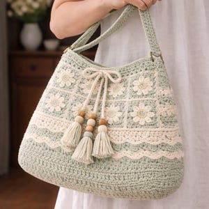 Puede incluir: Bolso tote de ganchillo en verde suave y crema. El bolso presenta un patrón floral, borlas decorativas y cuentas de madera. Tiene una correa para el hombro y lo sostiene una persona.