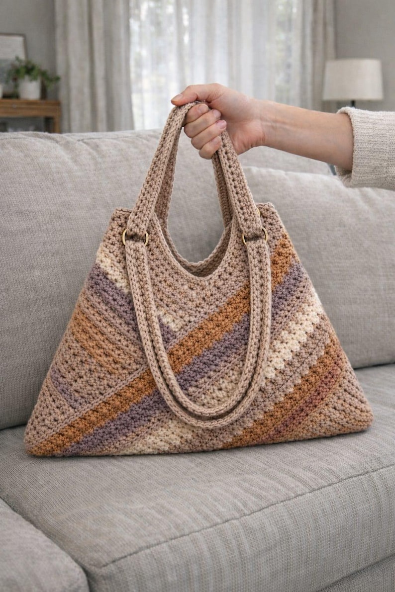 以下が含まれることがあります： ベージュ、茶色、紫の色合いのストライプ模様が特徴の、手作りのかぎ針編みトートバッグ。三角形の形状で、2つのハンドルと金色のリングが付いています。グレーのソファの上に置かれています。