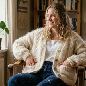 Peut inclure: Un cardigan en tricot crème épais avec de gros boutons en bois et des poches avant. Le cardigan est porté sur un haut blanc et associé à un jean bleu. La personne est assise dans une chaise en bois, souriante, avec des plantes en arrière-plan.