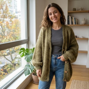 Peut inclure: Une femme porte un cardigan vert olive boutonné avec des boutons en bois, un haut gris et un jean bleu. Le cardigan a une texture. Elle sourit devant une fenêtre avec une plante.