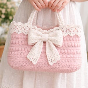 May include: A pink crochet handbag with a large cream-colored bow and matching trim. The bag has a textured pattern and sturdy handles. The bag is held by a person wearing a white dress.