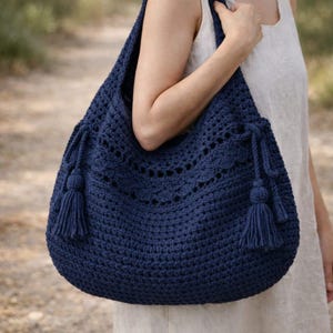 Könnte beinhalten: Eine marineblaue Häkeltasche mit einer großen, abgerundeten Form. Die Tasche hat dekorative Lochmuster und Quasten auf jeder Seite. Die Tasche wird über der Schulter getragen.