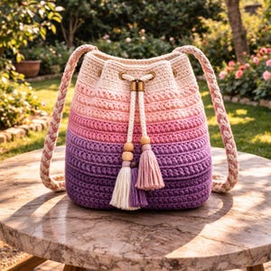 Könnte beinhalten: Handgefertigte Häkeltasche in Farbverlauf von Creme über Rosa bis Lila. Die Tasche hat geflochtene Träger, einen Kordelzugverschluss und dekorative Quasten. Die Tasche steht auf einem Marmortisch.