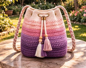 May include: A handmade crochet bucket bag in a gradient of colors, from cream to pink and purple. The bag features braided straps, a drawstring closure, and decorative tassels. The bag is sitting on a marble table.