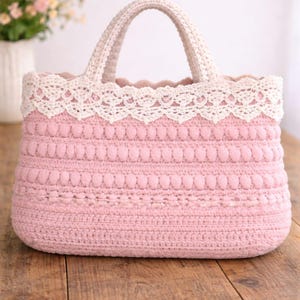 Pu&ograve; includere: Borsa tote rosa all'uncinetto fatta a mano con finiture e manici in pizzo color crema. La borsa presenta un design strutturato con file di bobble e una base rettangolare. La borsa &egrave; appoggiata su una superficie di legno.