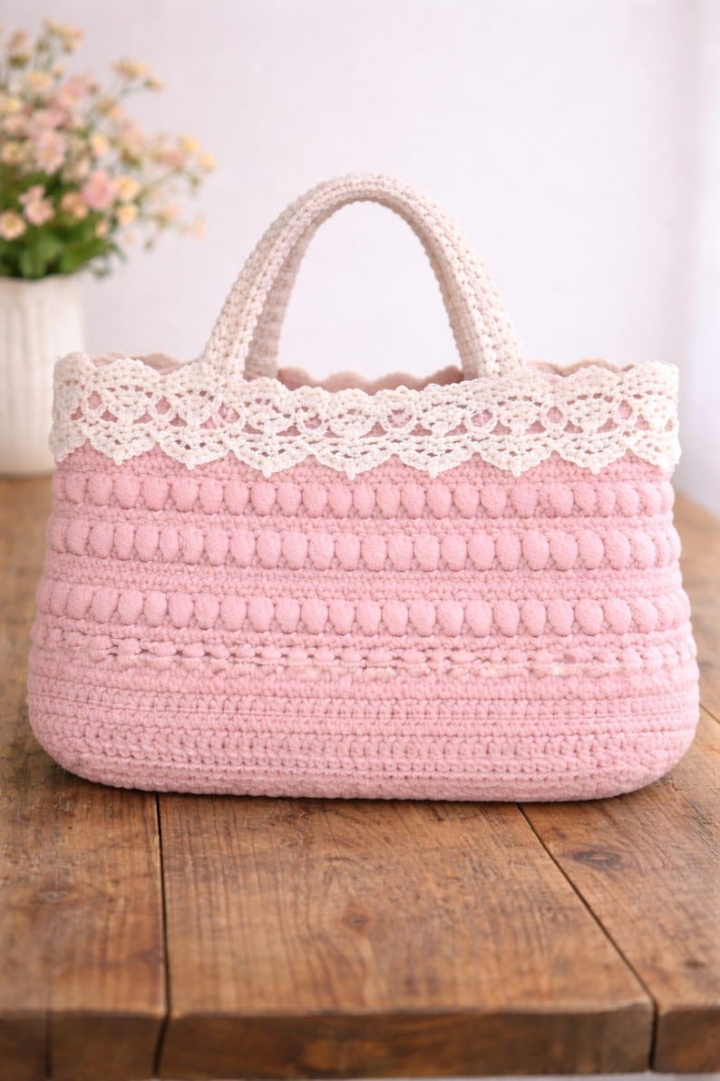 Pu&ograve; includere: Borsa tote rosa all'uncinetto fatta a mano con finiture e manici in pizzo color crema. La borsa presenta un design strutturato con file di bobble e una base rettangolare. La borsa &egrave; appoggiata su una superficie di legno.