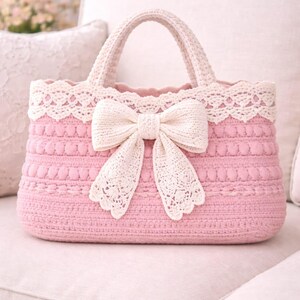 Pu&ograve; includere: Borsa a mano rosa all'uncinetto con finiture in pizzo color crema e un grande fiocco. La borsa ha una forma strutturata con un manico ed &egrave; appoggiata su una superficie chiara. La borsa &egrave; larga circa 30 cm.