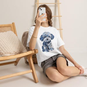 May include: A white short-sleeved t-shirt featuring a graphic print of a puppy composed of various shades of blue denim fabric patches. The puppy design displays a textured, patchwork appearance with visible stitching details throughout the dog.