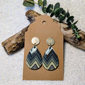 Może przedstawiać: Kolczyki w kształcie łezki ze złotymi krążkami i wzorem w szewrony w kolorach czarnym, białym i zielonym. Kolczyki są prezentowane na brązowej etykiecie, z kawałkiem drewna i zielenią w tle. Stylowy dodatek.