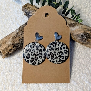 Può includere: Un paio di orecchini rotondi con stampa leopardata e top a forma di cuore argentati. Gli orecchini sono esposti su una carta marrone con un foro nella parte superiore, su uno sfondo bianco strutturato e un pezzo di legno alla deriva.