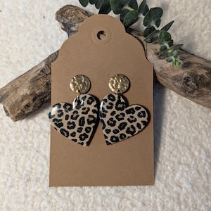 May include: A pair of heart-shaped earrings with a leopard print design. The earrings have a tan background with black spots and are topped with gold-colored, hammered-texture circles. The earrings are displayed on a brown tag.