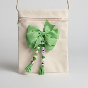 May include: A cream-coloured crossbody bag with a green bow and beaded tassel. The bag has a zip closure and a long strap. The tassel has the name "JOEY" spelled out in letter beads. The bow and tassels are a bright green colour.