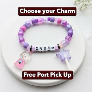 Könnte beinhalten: Ein Armband aus lila, rosa und weißen Perlen, mit dem Wort "DREAM" in Buchstabenperlen geschrieben. Es enthält Anhänger wie einen Kaugummiautomaten und einen sternförmigen Lutscher. Der Text "Choose your Charm" und "Free Port Pick Up" sind ebenfalls sichtbar.