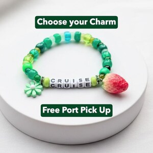 May include: A beaded bracelet with green, blue, and white beads. The bracelet has the word "CRUISE" spelled out in white letter beads. It features a strawberry charm and a green flower charm. The text "Choose your Charm" and "Free Port Pick Up" are also visible.