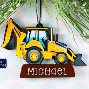 May include: A yellow and black wooden excavator ornament with the name "MICHAEL" in silver lettering. The ornament is hanging from a black string and is decorated with green pine needles.
