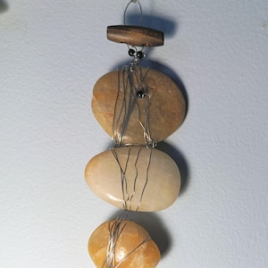 May include: A handcrafted mobile featuring three smooth, oval stones in shades of tan and cream, wrapped with silver wire. A small wooden bead and black beads are at the top, with a decorative silver button at the bottom.