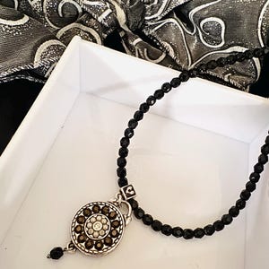 May include: A black beaded necklace with a silver pendant. The pendant has a floral design of brown and clear stones. The necklace is displayed in a white box with a black and white patterned scarf in the background.