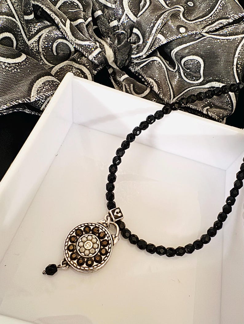 May include: A black beaded necklace with a silver pendant. The pendant has a floral design of brown and clear stones. The necklace is displayed in a white box with a black and white patterned scarf in the background.