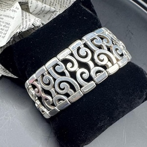 May include: A silver-toned bracelet with an openwork design of swirling scroll patterns. The bracelet is resting on a black velvet surface, showcasing its intricate details and craftsmanship. A stylish accessory.