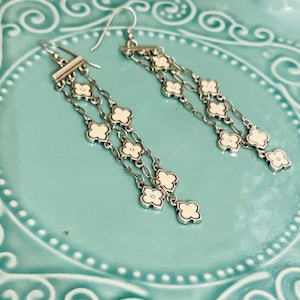 May include: A pair of silver-toned dangle earrings featuring multiple delicate chains suspended from a horizontal bar. Each chain is adorned with small, four-petaled clover-shaped charms filled with a smooth, off-white enamel inlay.