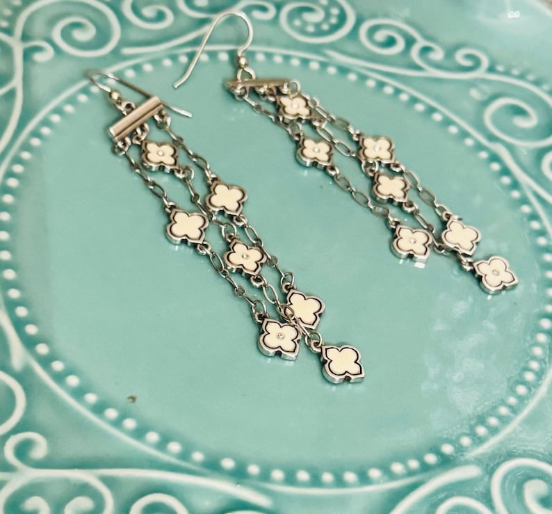May include: A pair of silver-toned dangle earrings featuring multiple delicate chains suspended from a horizontal bar. Each chain is adorned with small, four-petaled clover-shaped charms filled with a smooth, off-white enamel inlay.